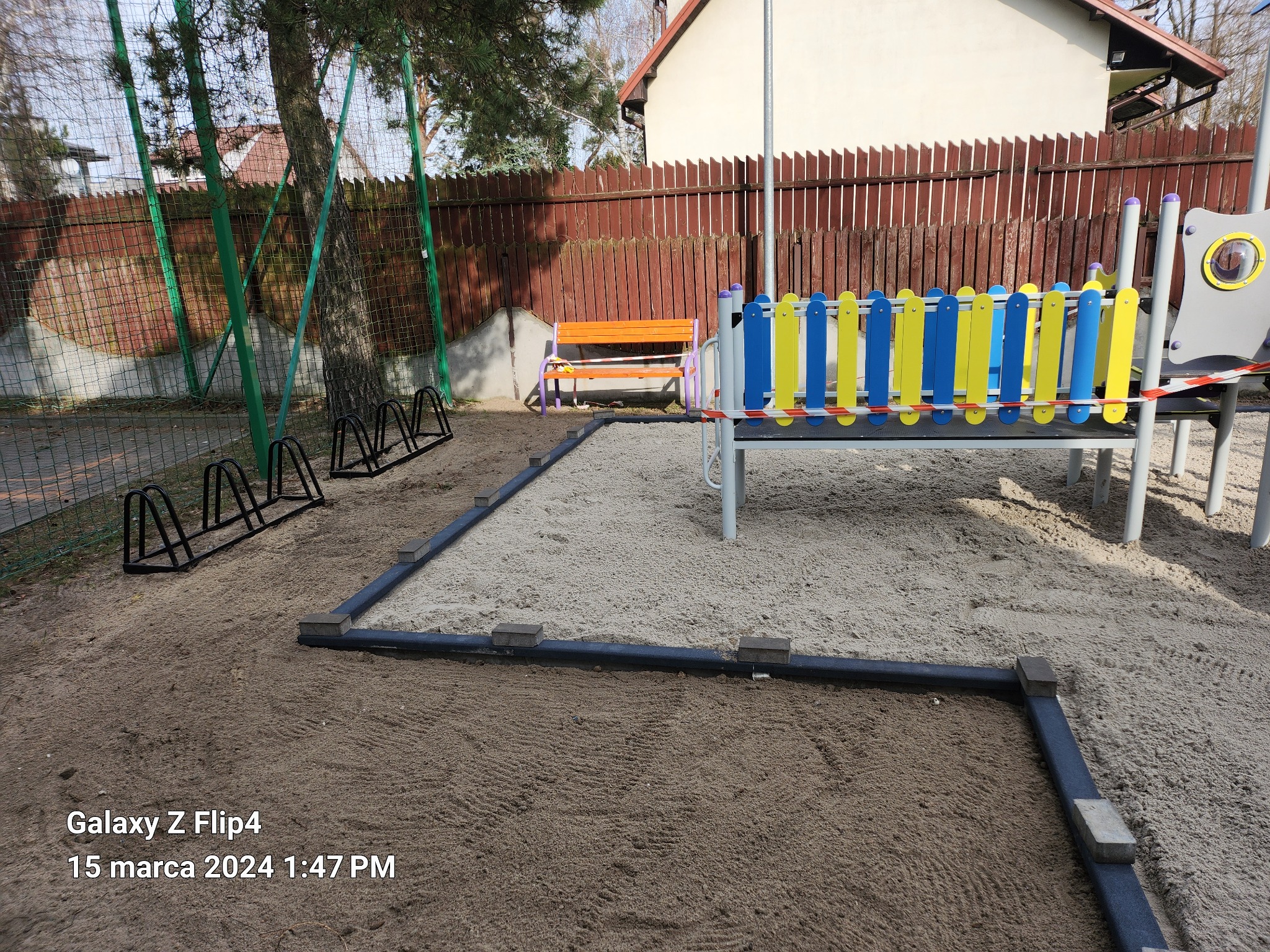 Przygotowany teren pod plac zabaw z obrzeżami i piaskiem, widoczny stojak na rowery, ławka i elementy zabawowe w tle.