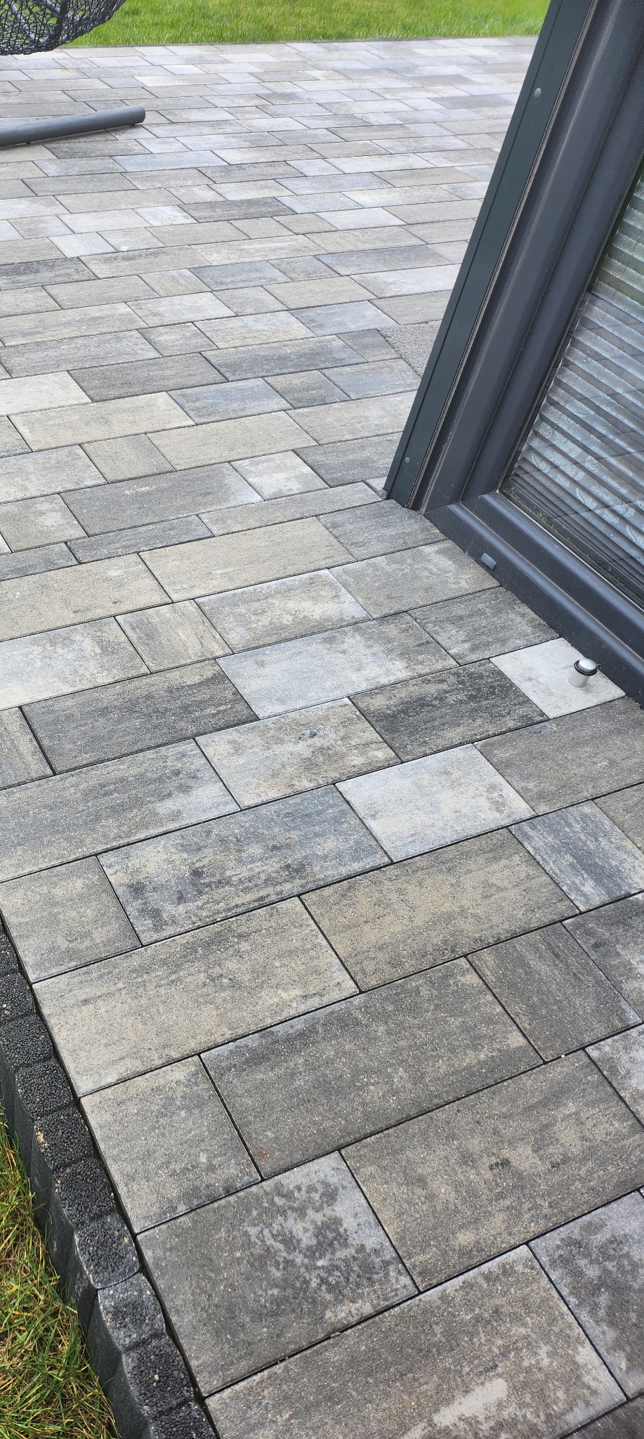 Ułożona szara kostka brukowa o prostokątnym kształcie przylegająca do ciemnej ramy okiennej i obrzeża z kostki brukowej, widoczne źdźbła trawy.