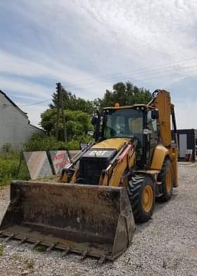 Żółta koparko-ładowarka marki CAT z brudną łyżką na żwirowym podłożu, widoczne trójkątne tablice ostrzegawcze i budynek w tle.
