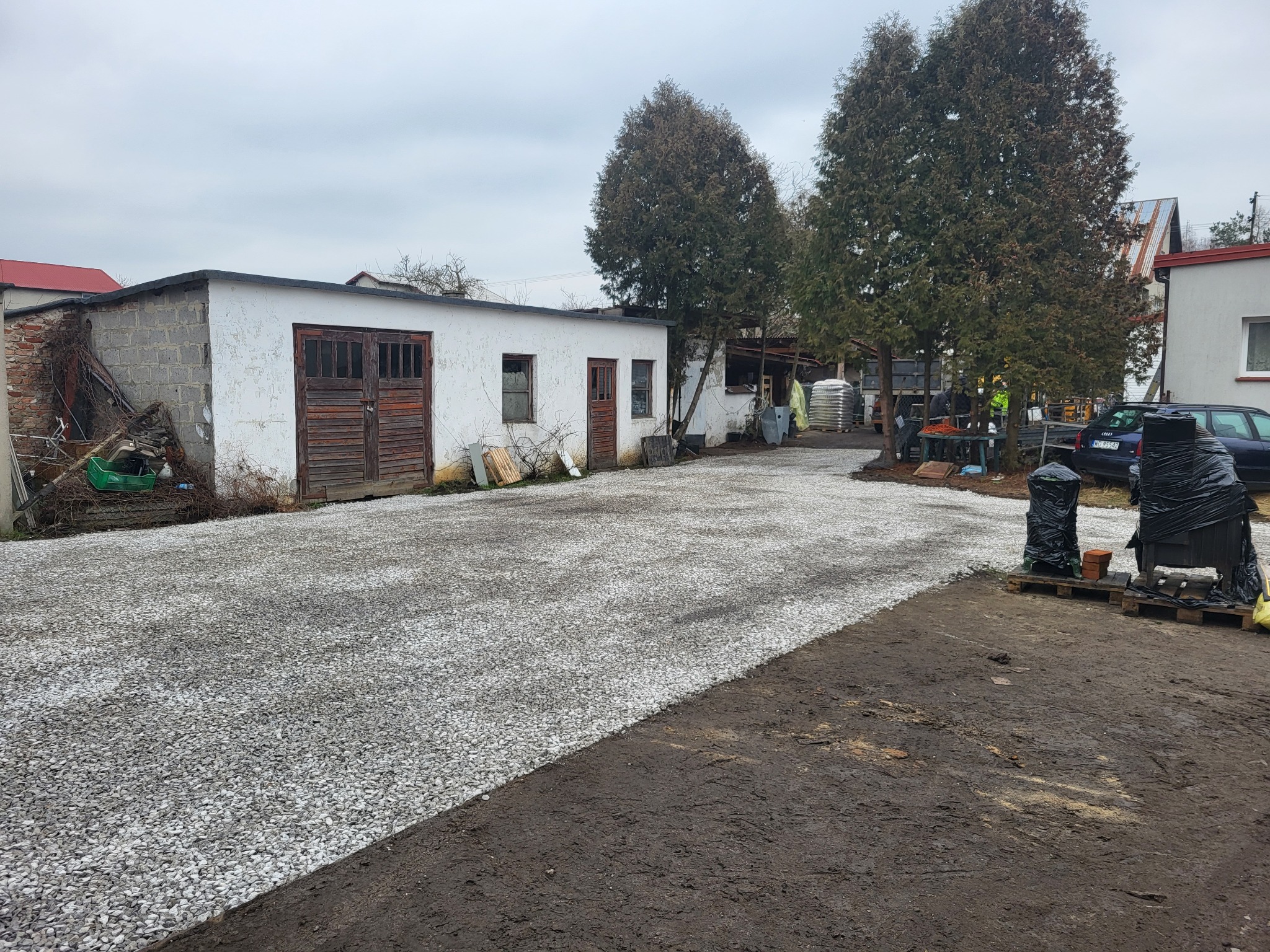 Podwórko z budynkami gospodarczymi i nowo utwardzoną drogą żwirową, widoczne drzewa, samochód osobowy i elementy budowlane zabezpieczone folią.