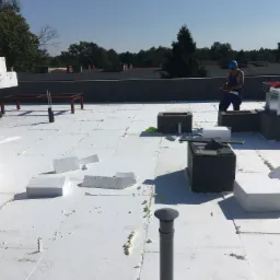 Montaż izolacji termicznej dachu płaskiego z płyt styropianowych, widoczny pracownik w niebieskim kombinezonie i kasku, oraz puszki z pianką montażową i elementy wentylacyjne.