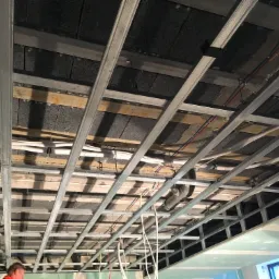 Metalowa konstrukcja sufitu podwieszanego z widocznymi instalacjami: rury wentylacyjne, kable elektryczne, oraz częściowo zamontowane płyty gipsowo-kartonowe.