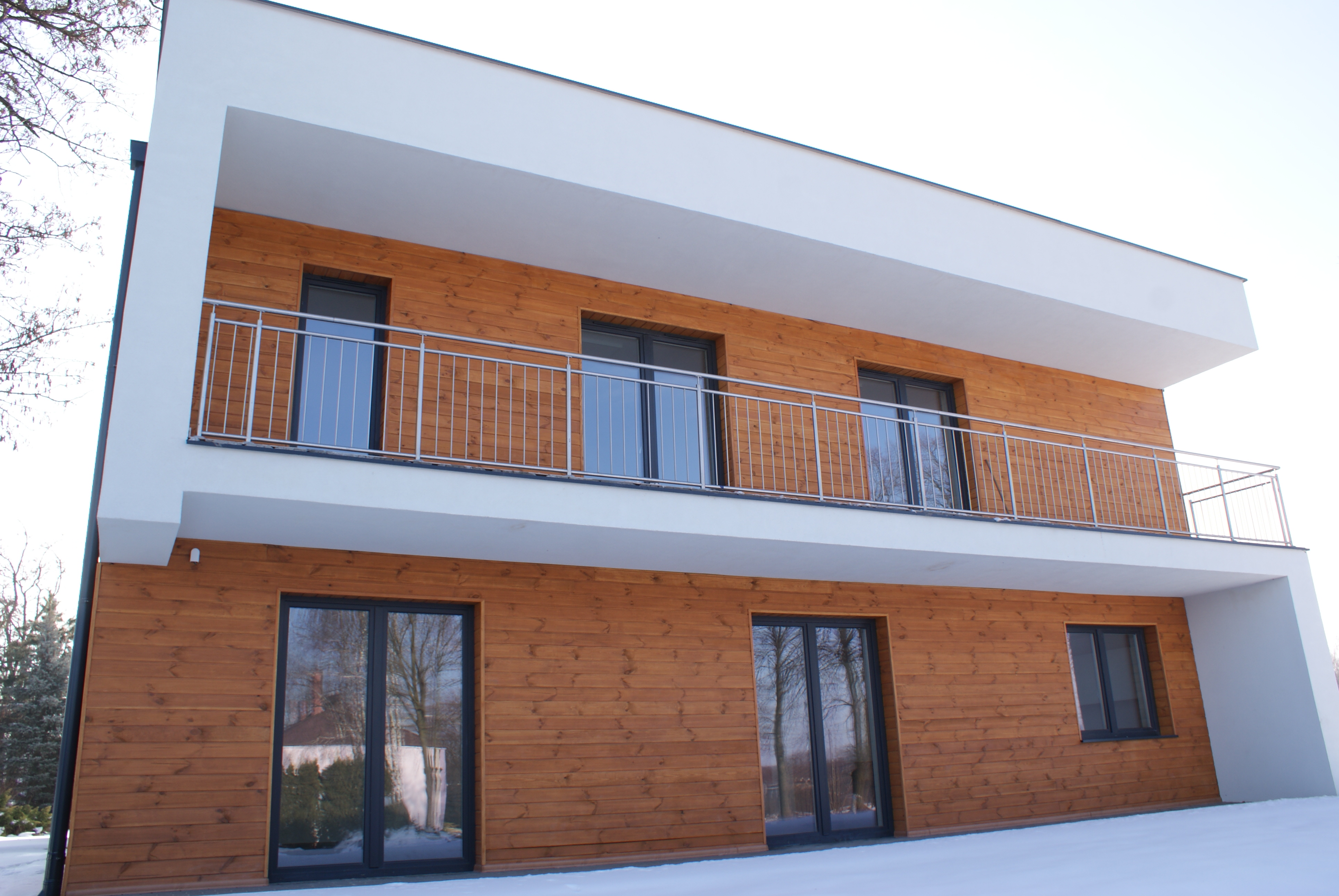 Nowoczesny, trzypiętrowy dom w Łodzi z drewnianą elewacją i białymi elementami architektonicznymi, widoczny z zewnątrz w zimowej scenerii.