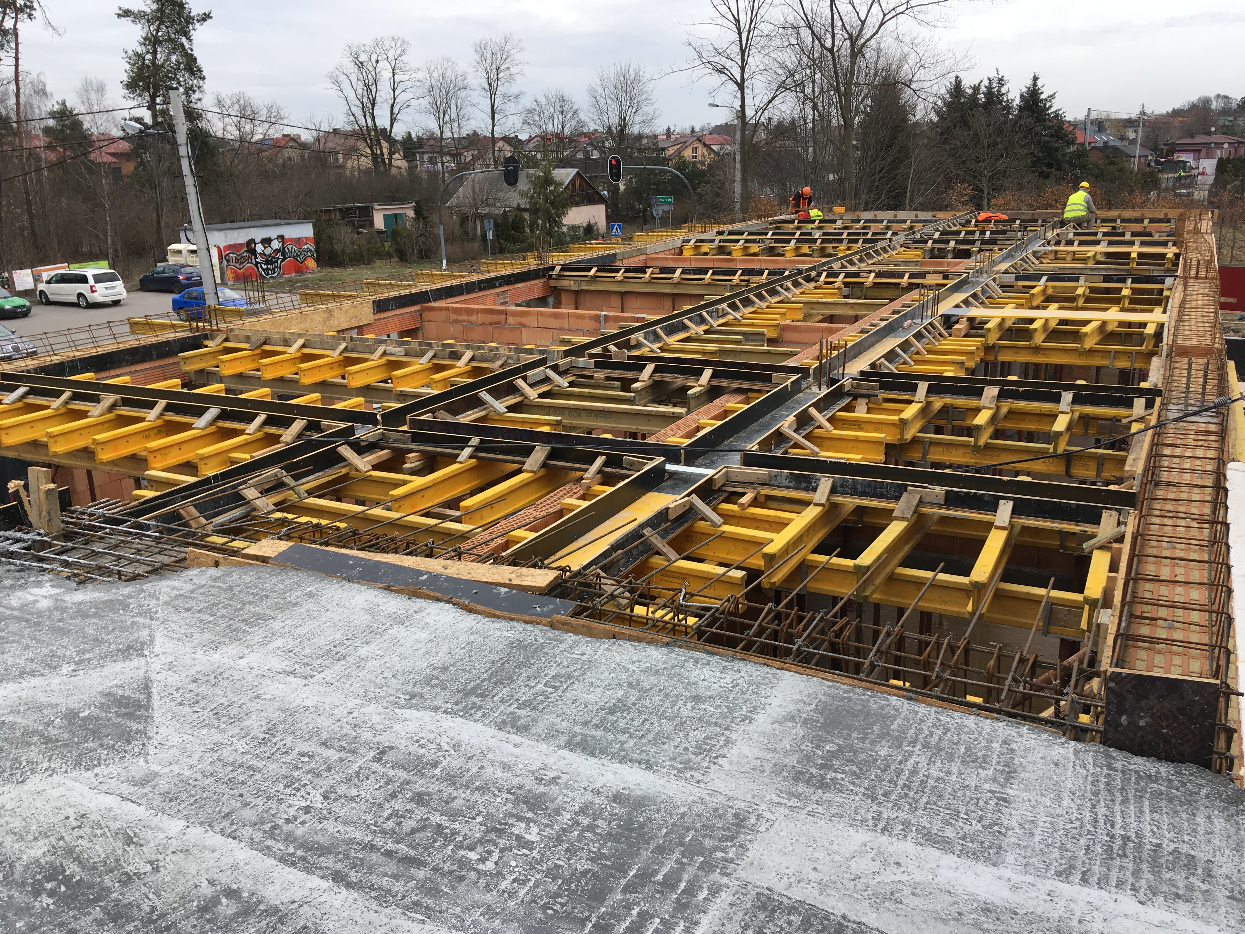 Zbrojenie i szalunek pod płytę fundamentową z widocznymi elementami drewnianymi i stalowymi, robotnicy w odblaskowych kamizelkach w tle, widok z góry na plac budowy z elementami infrastruktury...