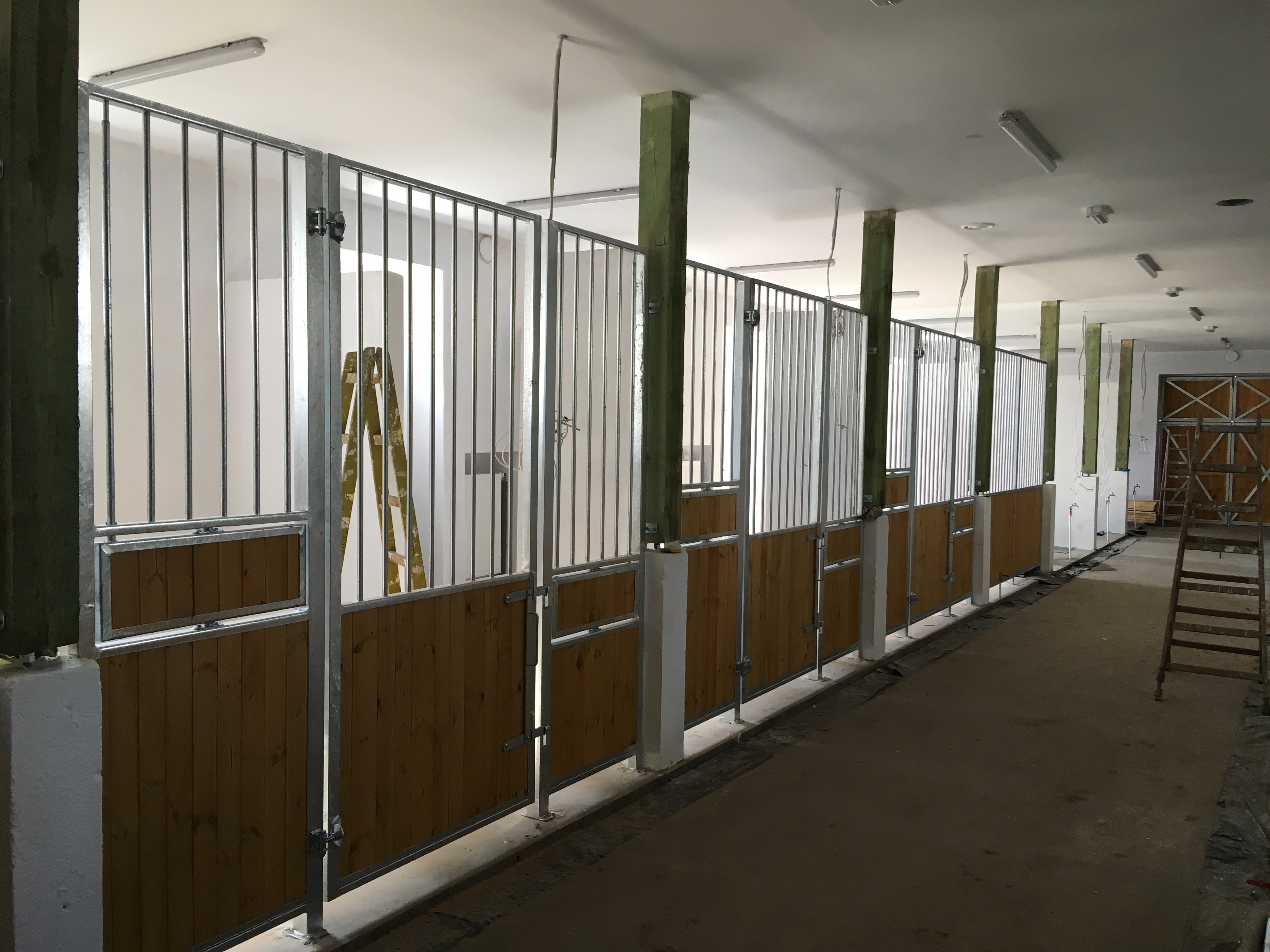 Wnętrze stajni z boksami dla koni w trakcie budowy, widoczne metalowe konstrukcje z drewnianymi panelami, drabina oparta o ścianę, surowy beton na podłodze i drewniane słupy podtrzymujące strop.