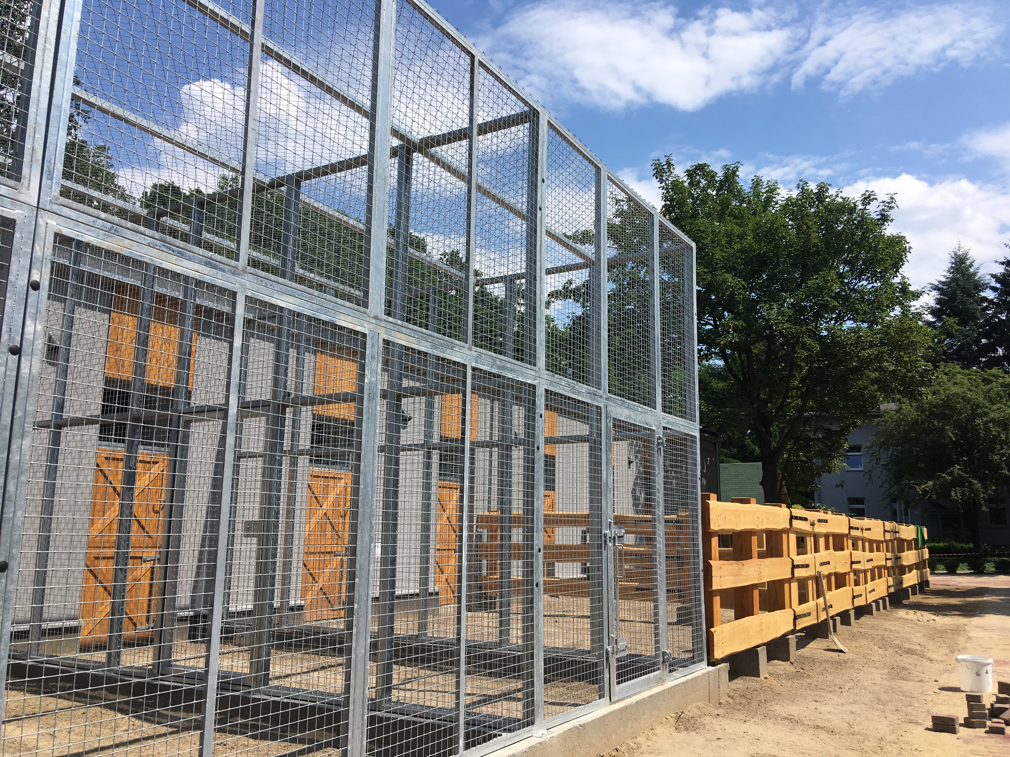 Konstrukcja z siatki stalowej z drewnianymi elementami, prawdopodobnie klatka dla zwierząt lub woliera, z widocznymi drzwiami i tłem nieba oraz drzew