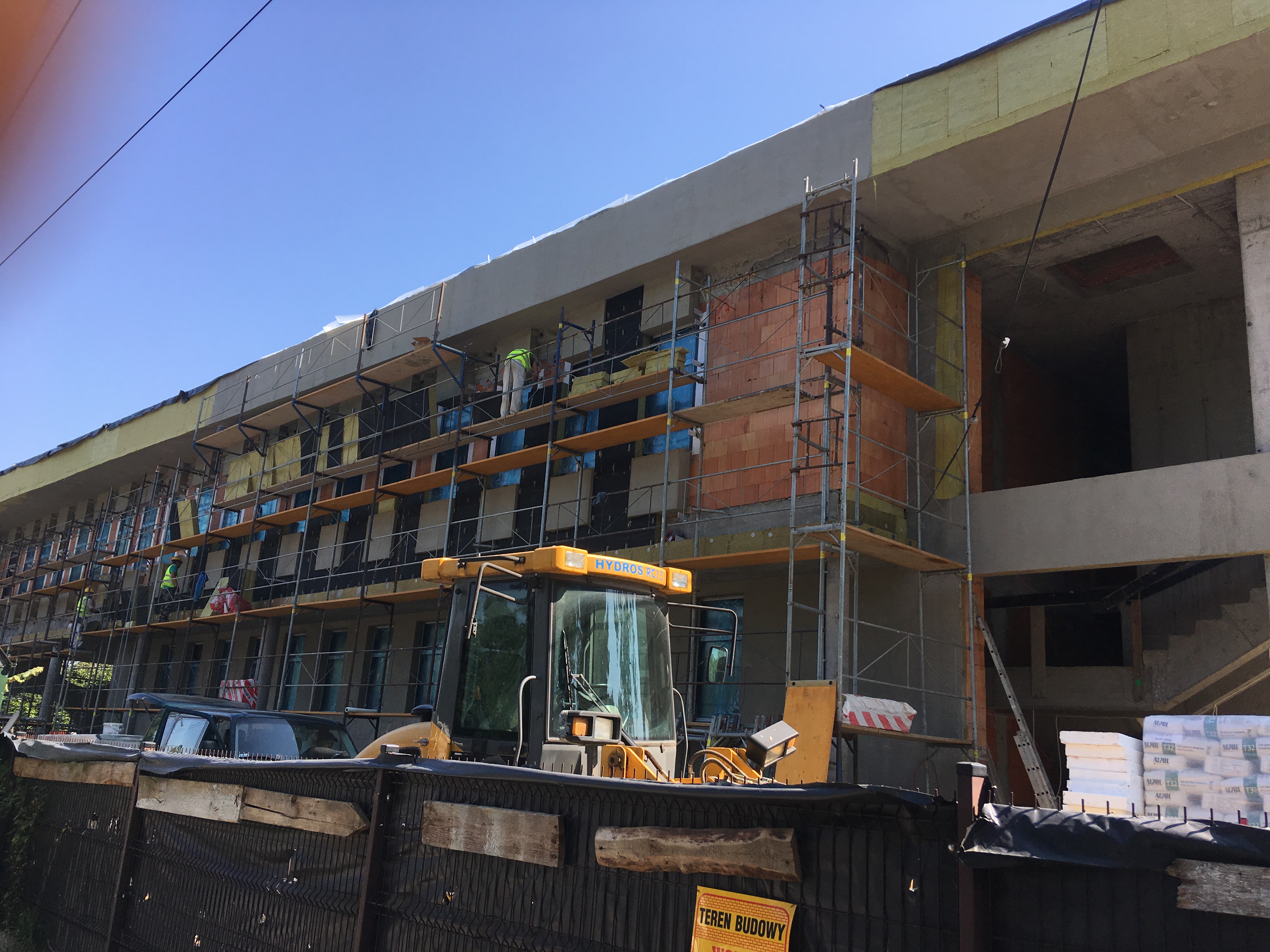 Wielopiętrowy budynek w trakcie budowy, z rusztowaniami, widocznymi cegłami i ociepleniem, oraz żółtą maszyną budowlaną Hydros RC15 na pierwszym planie.