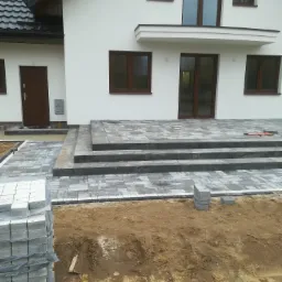 Etap budowy tarasu przed domem z szarej kostki brukowej. Widoczne ułożone schody i fragment tarasu, paleta z kostką oraz przygotowane podłoże.