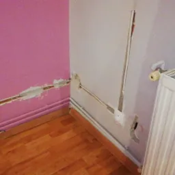 Wnętrze pokoju z uszkodzoną ścianą i widocznymi przewodami elektrycznymi, przygotowane do ukrycia instalacji w ścianie, obok grzejnik.