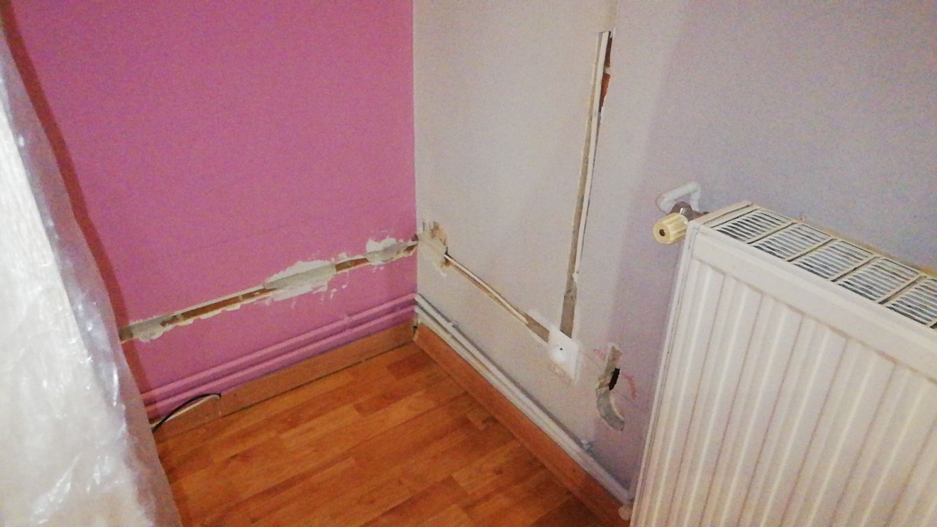 Wnętrze pokoju z uszkodzoną ścianą i widocznymi przewodami elektrycznymi, przygotowane do ukrycia instalacji w ścianie, obok grzejnik.