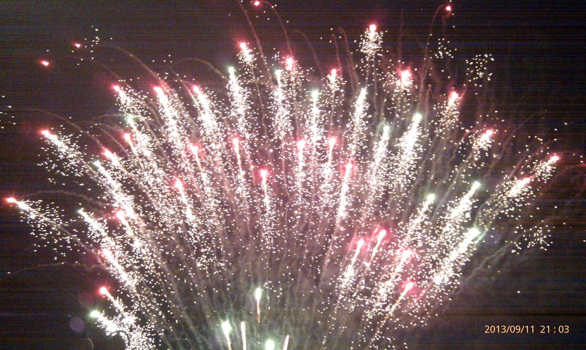 Wielobarwne fajerwerki rozświetlają nocne niebo, widoczne smugi światła i iskry w odcieniach bieli, czerwieni i zieleni, zdjęcie z datą w prawym dolnym rogu.