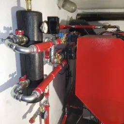 Czerwony piec CO z czarnym zasobnikiem, rurami w izolacji i manometrami w kotłowni, widok z dołu.