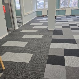 KAM-AN ANNA MITURA - Wnętrze biura z nową, wzorzystą wykładziną dywanową w odcieniach szarości. Widoczne okna, kolumna i fragment drzwi. Wykładzina ułożona w kwadraty i prostokąty.