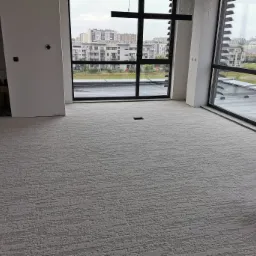 Jasnoszara, teksturowana posadzka jastrychowa w surowym biurze z dużymi oknami, widok na budynki mieszkalne w tle, wiadro z farbą w rogu.