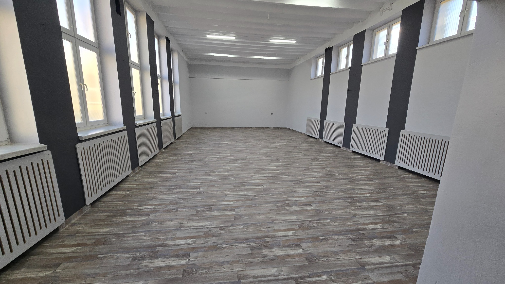 Jasne wnętrze po remoncie: panele podłogowe w odcieniach szarości, białe ściany z szarymi kolumnami, grzejniki z ażurowymi osłonami. Widok na całe pomieszczenie.