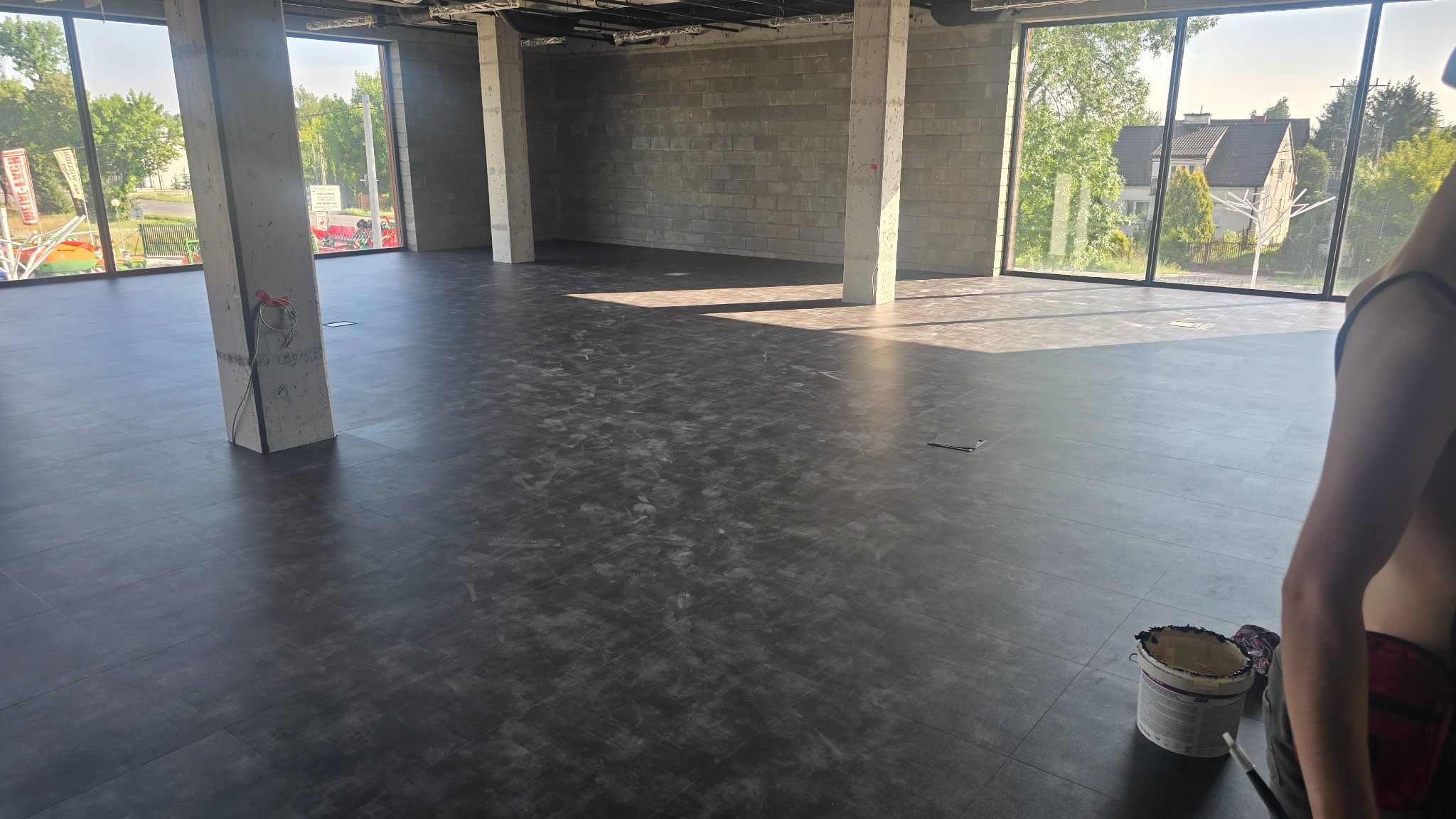 Nowa, ciemna podłoga w surowym biurze z betonowymi kolumnami i dużymi oknami. Widoczny fragment osoby i wiadro z farbą, sugerujące prace wykończeniowe.