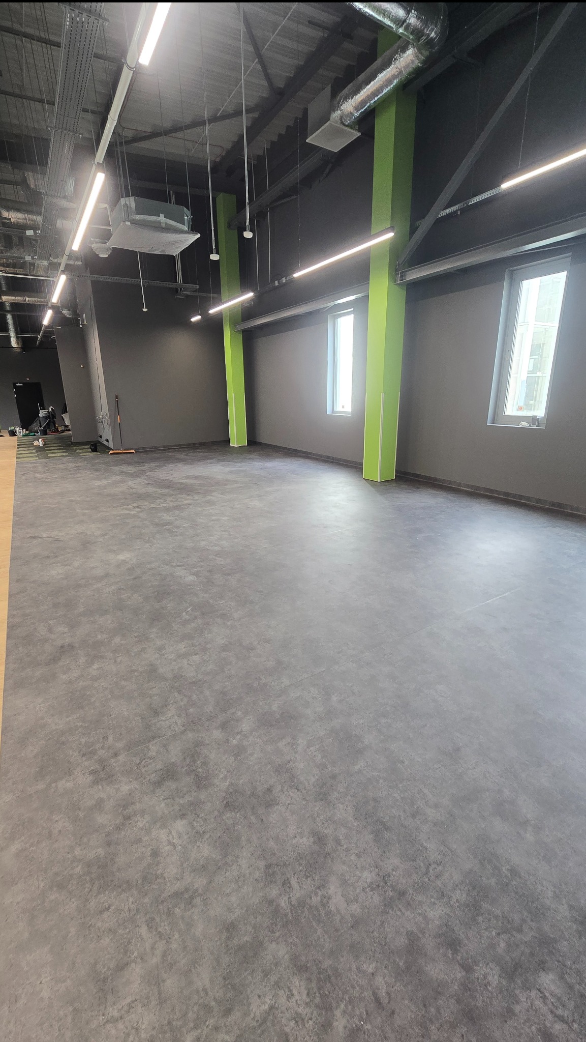 Szara, jednolita posadzka w przestronnym, jasnym wnętrzu biurowym z zielonymi kolumnami. Widoczne okna i instalacje pod sufitem. Częściowo widoczny sprzęt sprzątający.