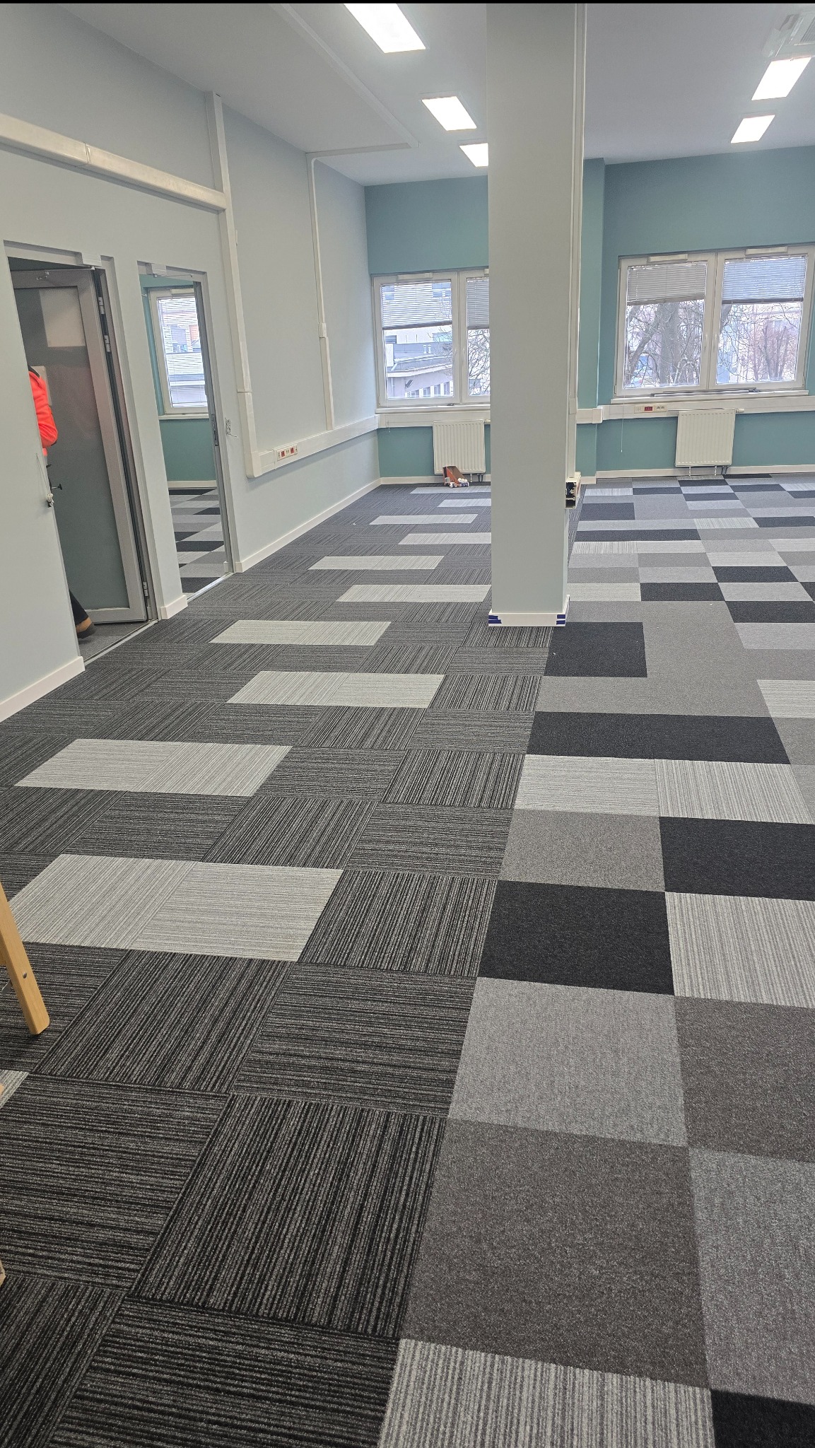Wnętrze biura z nową, wzorzystą wykładziną dywanową w odcieniach szarości. Widoczne okna, kolumna i fragment drzwi. Wykładzina ułożona w kwadraty i prostokąty.
