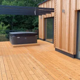 Drewniany taras z balustradą z czarną ramą i szklanym wypełnieniem, zadaszony pergolą, z jacuzzi na podwyższeniu, przylegający do domu z elewacją z jasnego drewna.