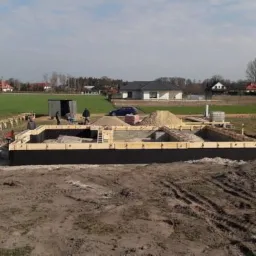 Fundamenty domu jednorodzinnego w trakcie budowy, widoczne szalunki, zbrojenie, zgromadzony piasek i cegły, w tle zabudowa wiejska i błękitne auto.