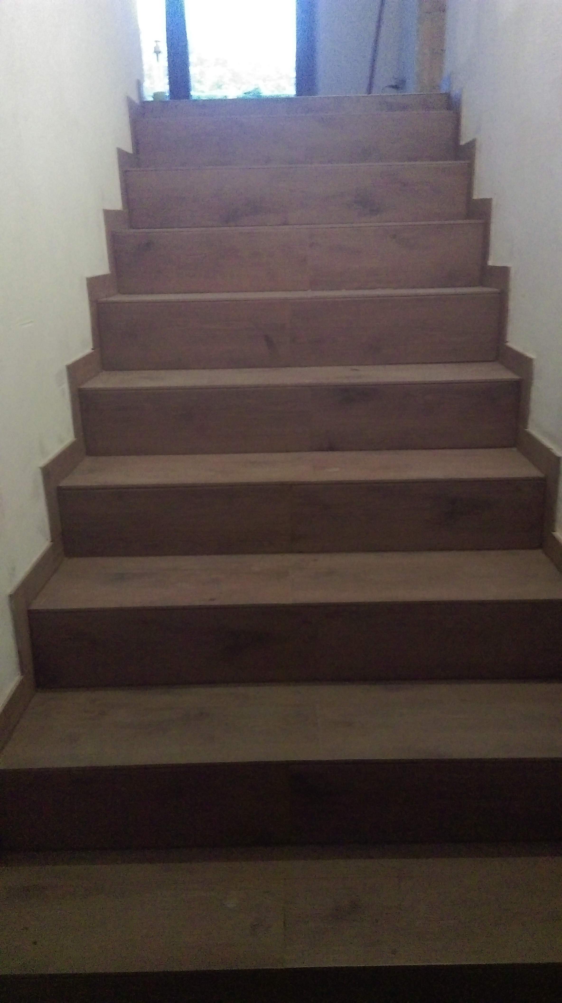 Schody wewnętrzne wyłożone panelami drewnopodobnymi, widok z dołu, jasne ściany, okno w tle.