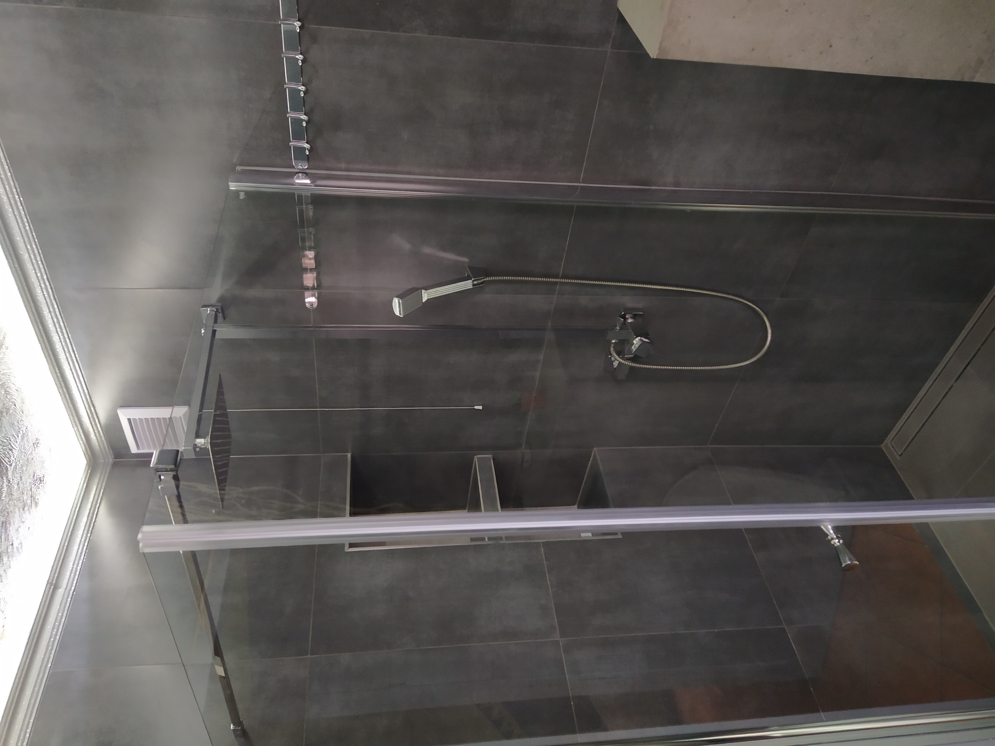 Nowoczesna kabina prysznicowa z ciemnoszarą glazurą, deszczownicą i odpływem liniowym, widok przez szkło kabiny.