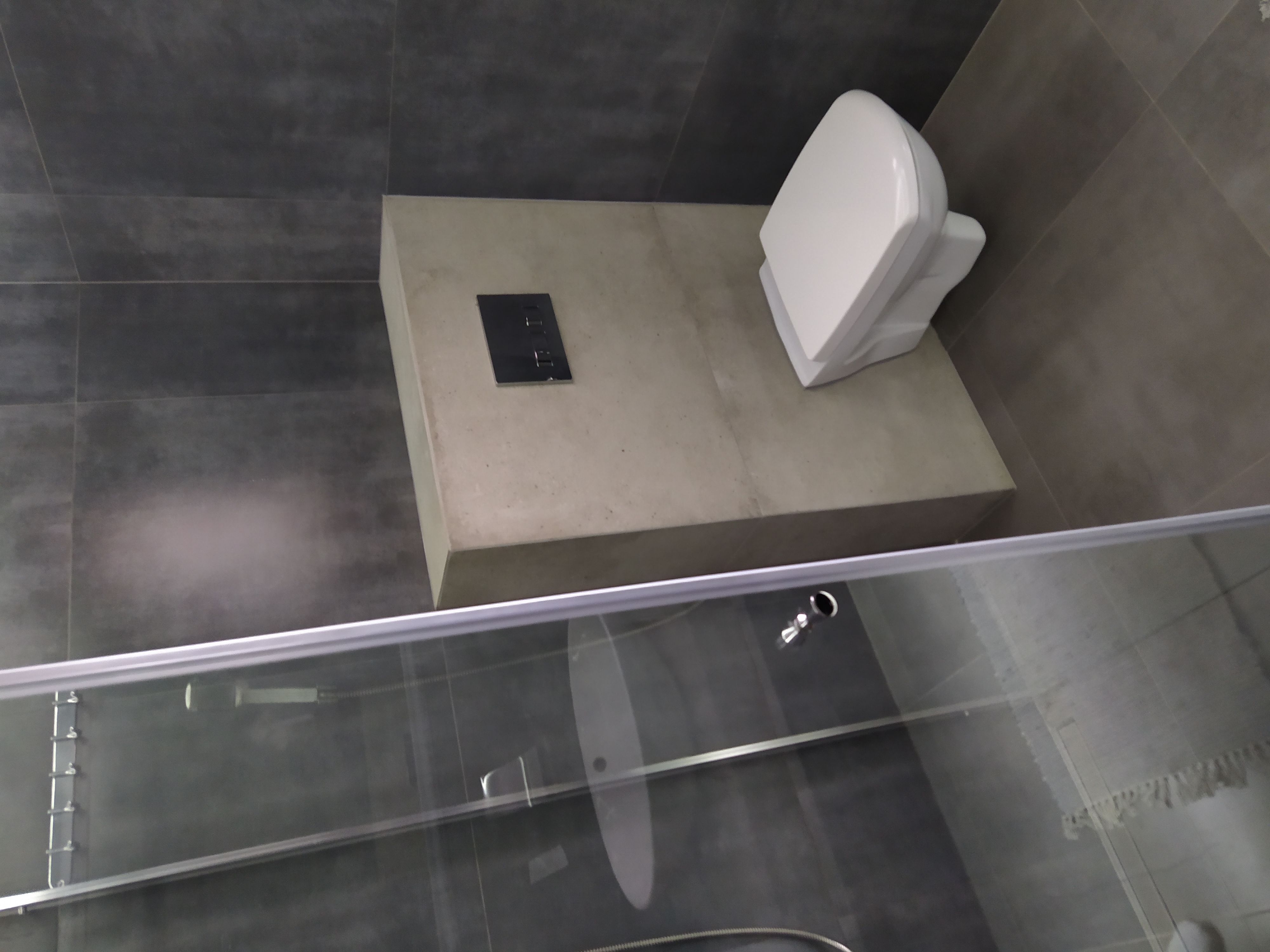 Nowoczesna toaleta podwieszana z szarą płytką na ścianach i podłodze, widok zza szklanej kabiny prysznicowej.