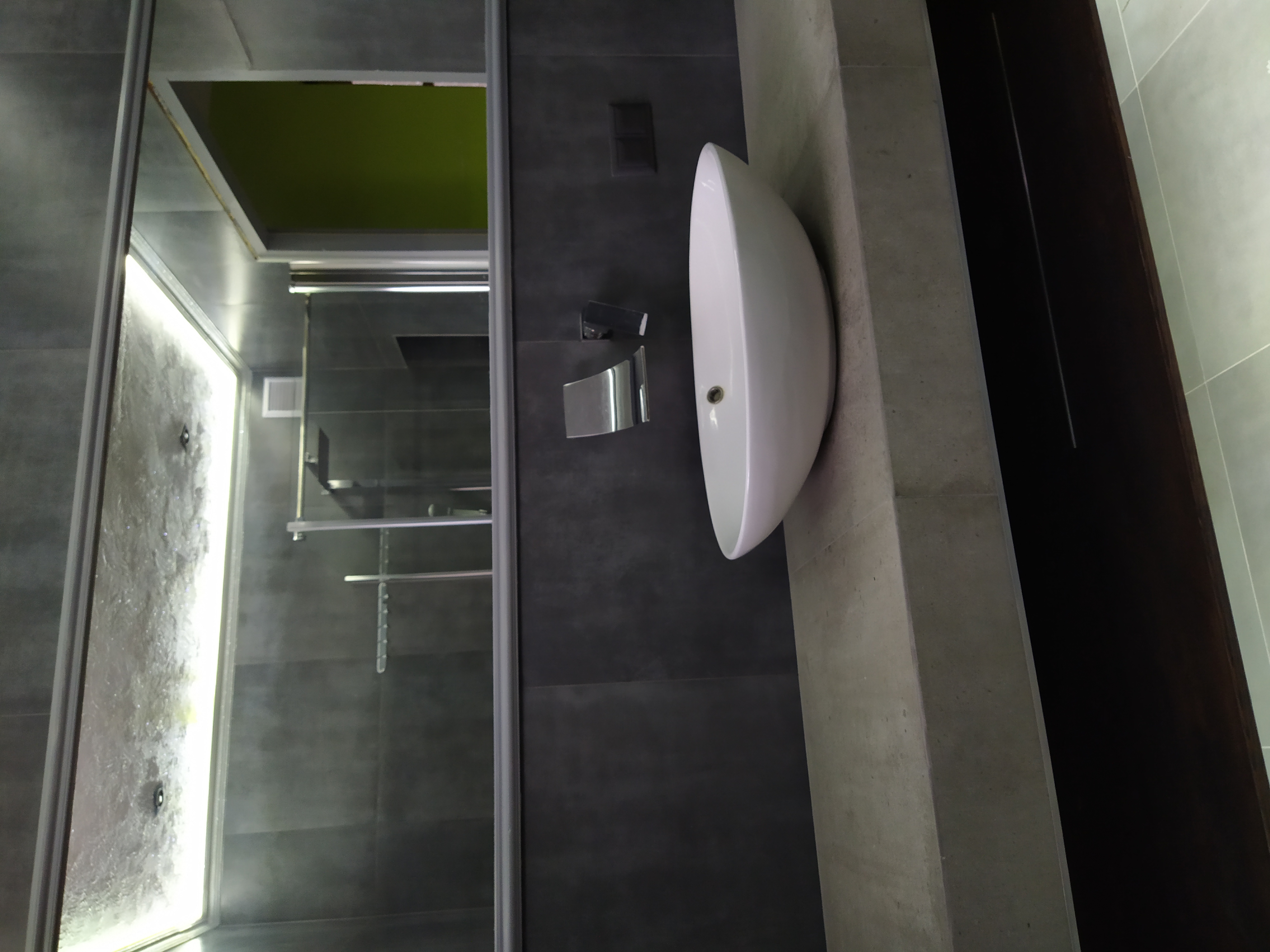 Nowoczesna łazienka z kabiną prysznicową z transparentnego szkła i owalną umywalką nablatową na tle ściany z ciemnoszarych płytek.