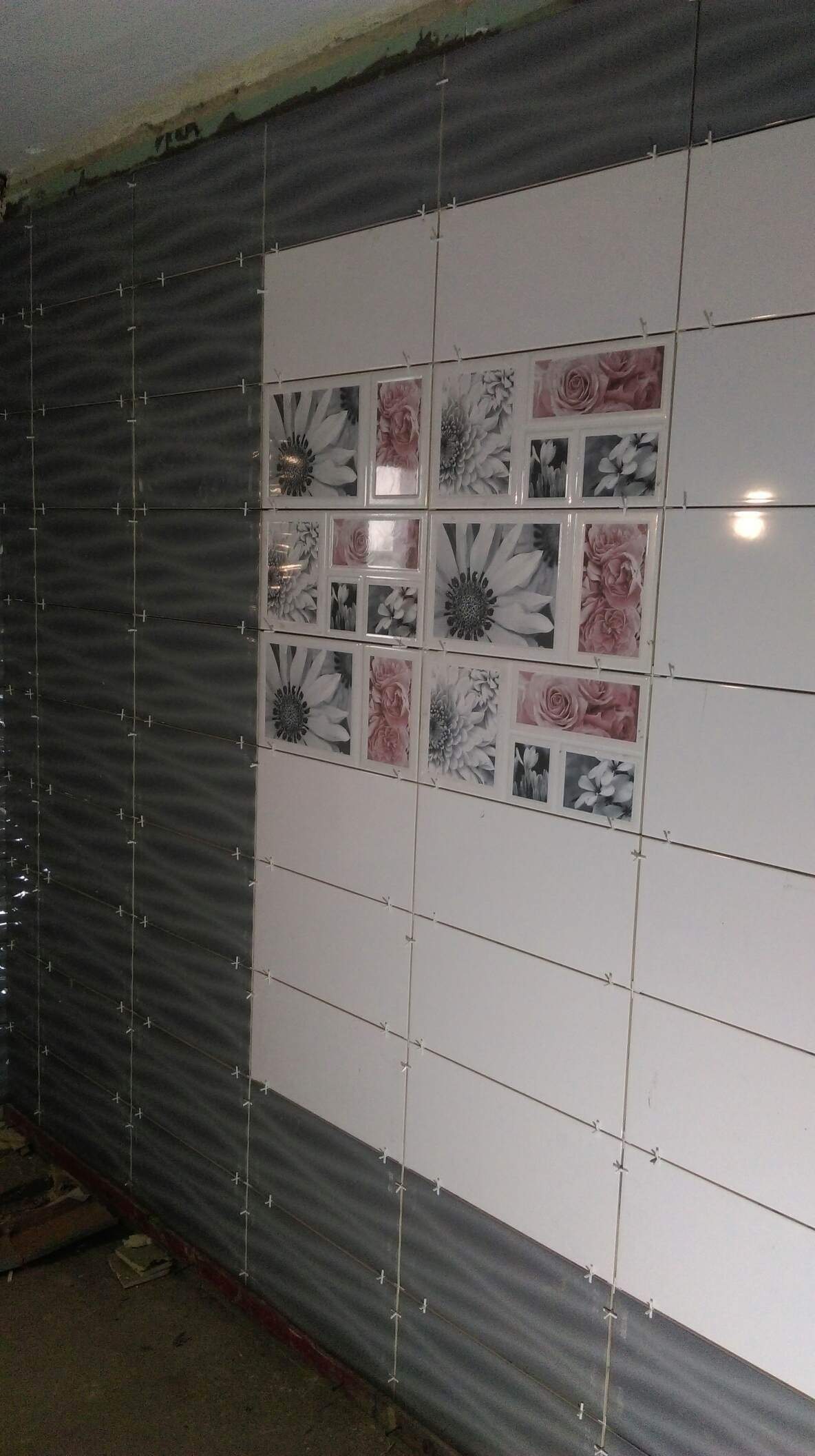 Ściana w trakcie układania płytek: szare płytki imitujące fale sąsiadują z białymi płytkami z motywem kwiatowym, widoczne krzyżyki dystansowe.