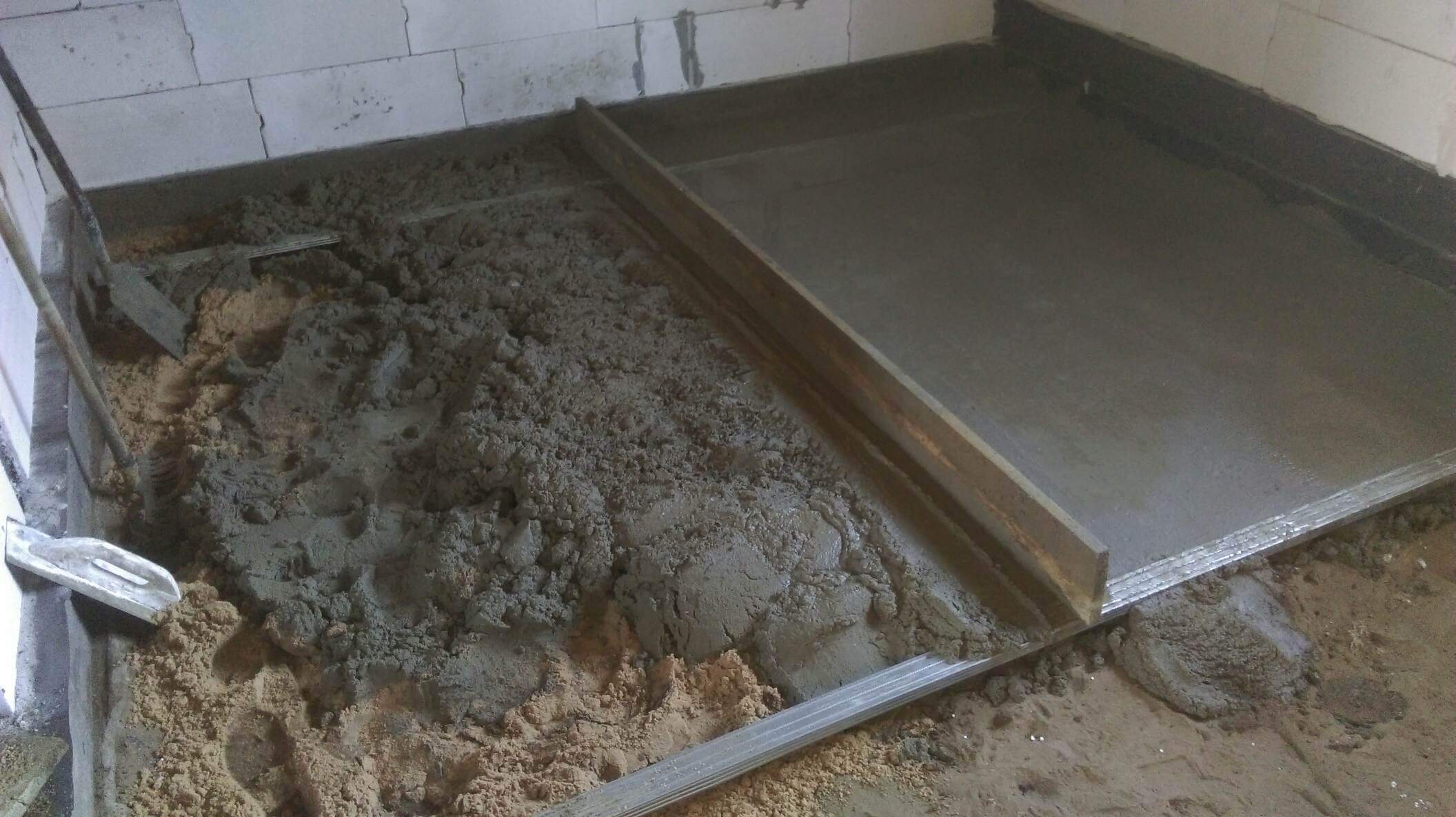 Wyrównywanie podłoża wylewką samopoziomującą, widoczne narzędzia: łata i kielnia, ściany z białego betonu komórkowego.