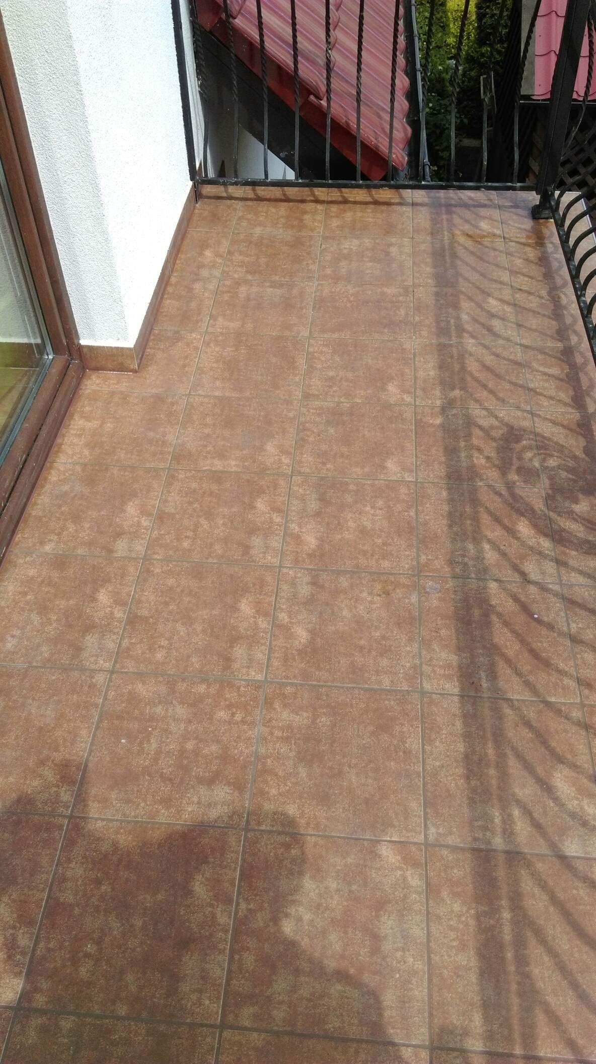 Balkon z brązowymi płytkami podłogowymi, widoczne cienie barierki i fragment ściany budynku.
