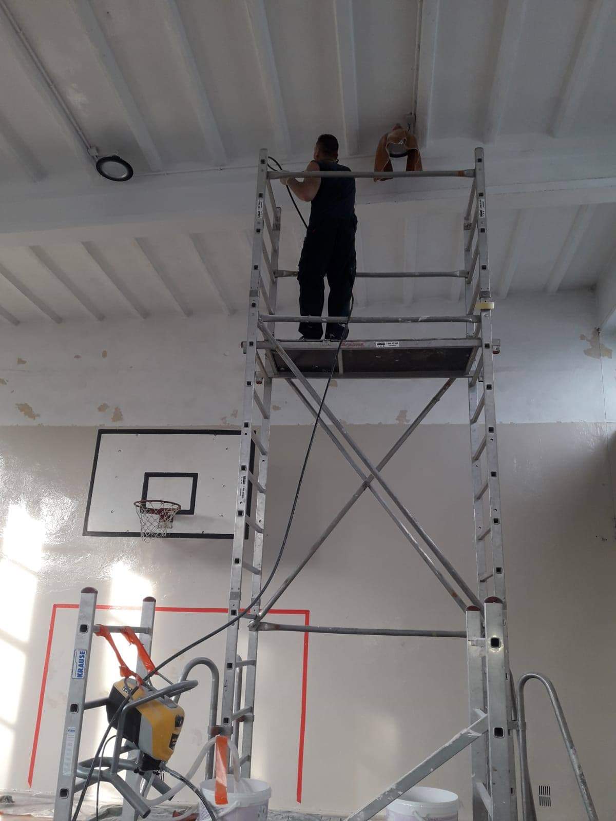 Pracownik na rusztowaniu maluje sufit w sali gimnastycznej, widoczny kosz do koszykówki i sprzęt malarski.