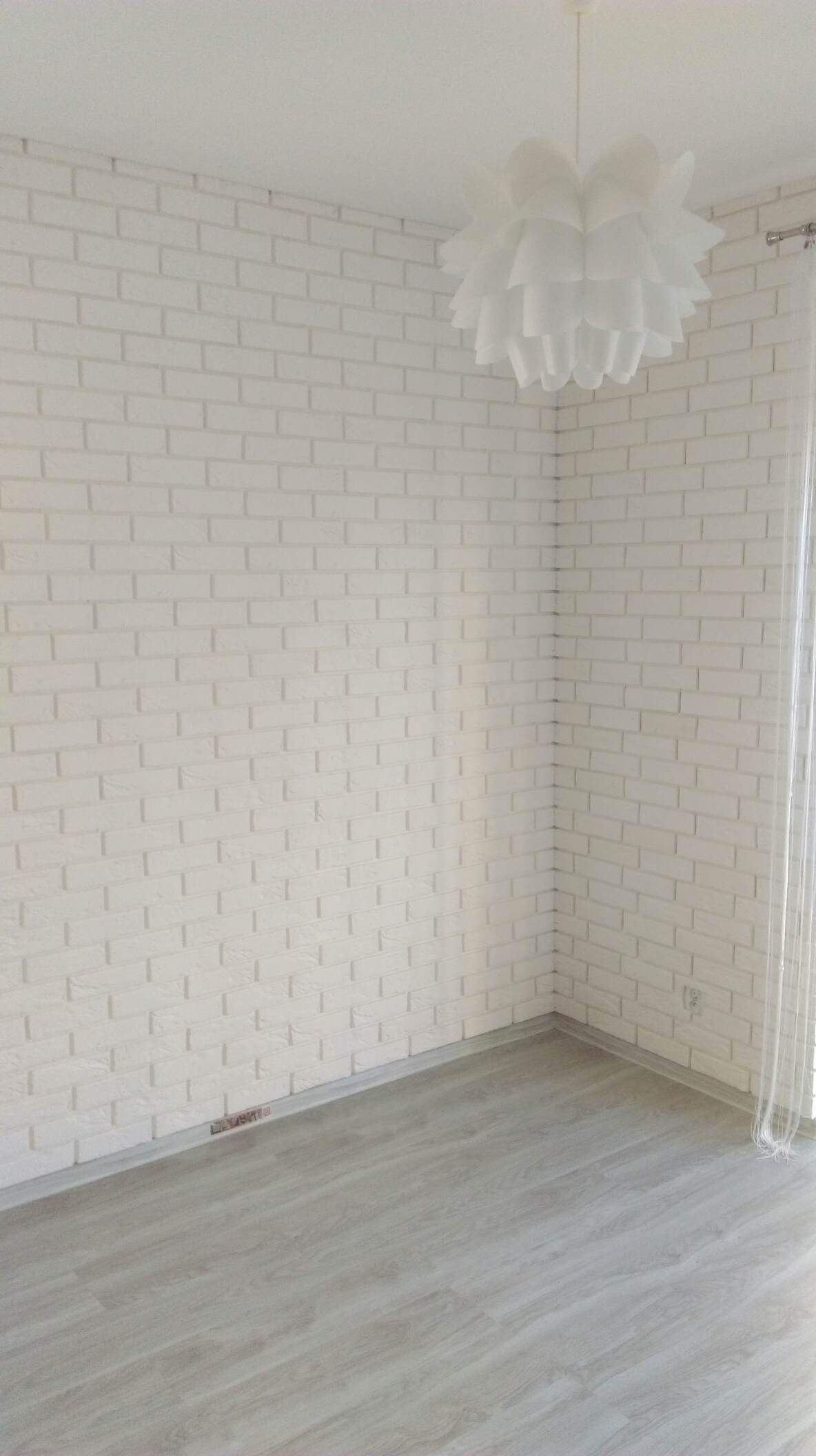 Jasne wnętrze pokoju z białą ścianą wyłożoną cegłami, szarą podłogą z paneli i dekoracyjną białą lampą wiszącą