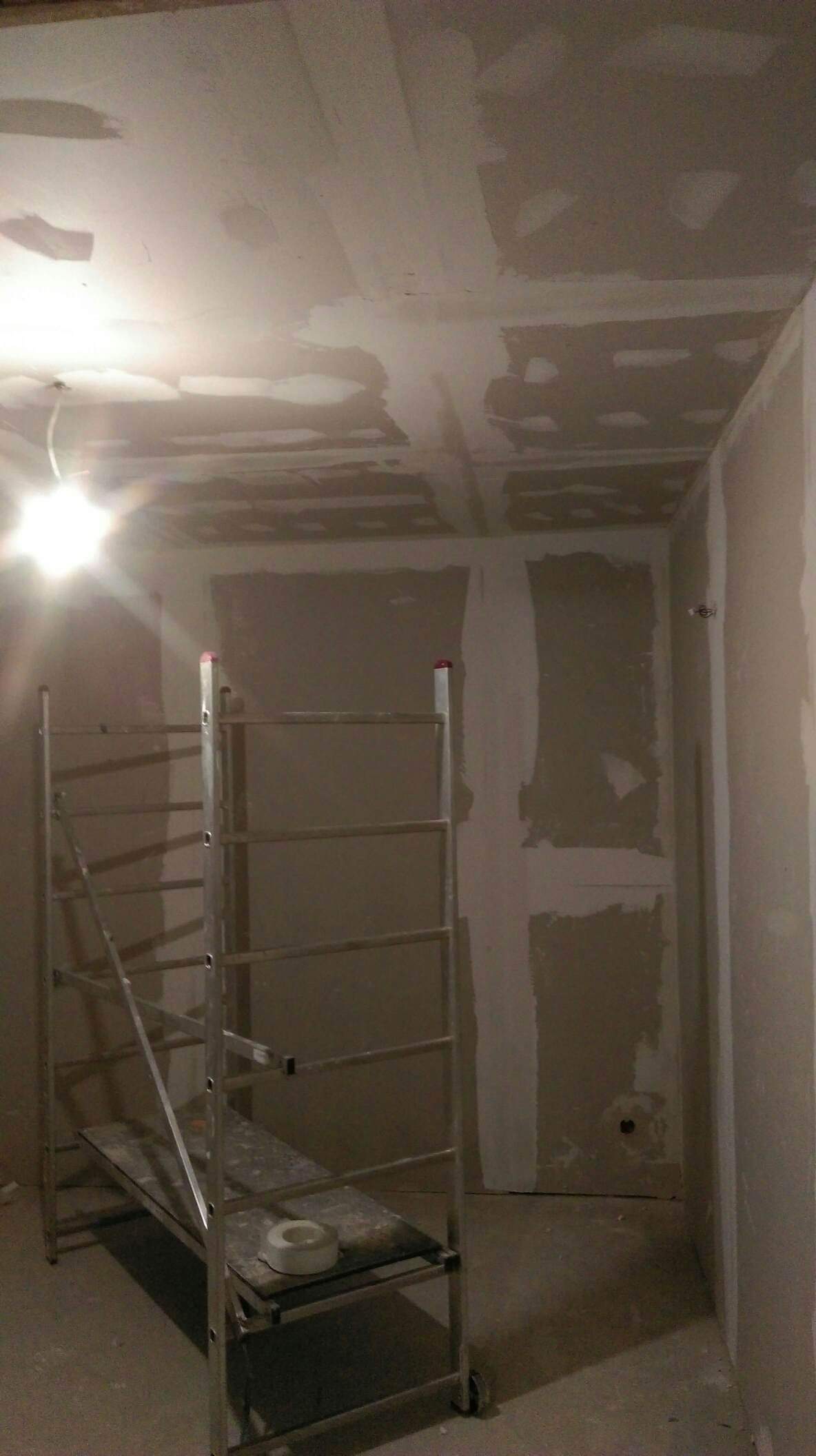 Wnętrze w trakcie prac przygotowawczych do malowania, widoczne ściany z nałożonymi łatami gipsowymi, aluminiowy ruszt na środku pomieszczenia, rolka taśmy malarskiej na podeście rusztowania, surowe...