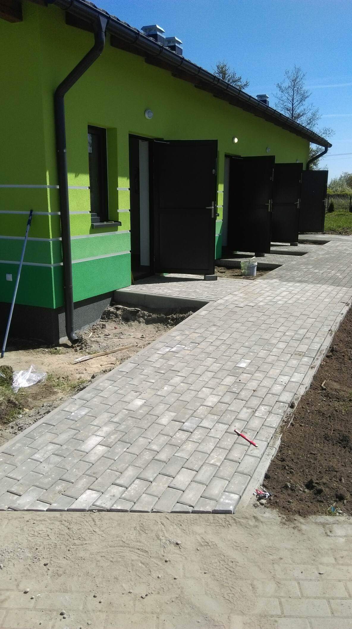 Ułożona szara kostka brukowa prowadząca do budynku z zieloną elewacją i czarnymi drzwiami, widoczny czerwony ołówek na kostce, ślady prac ziemnych po bokach ścieżki.