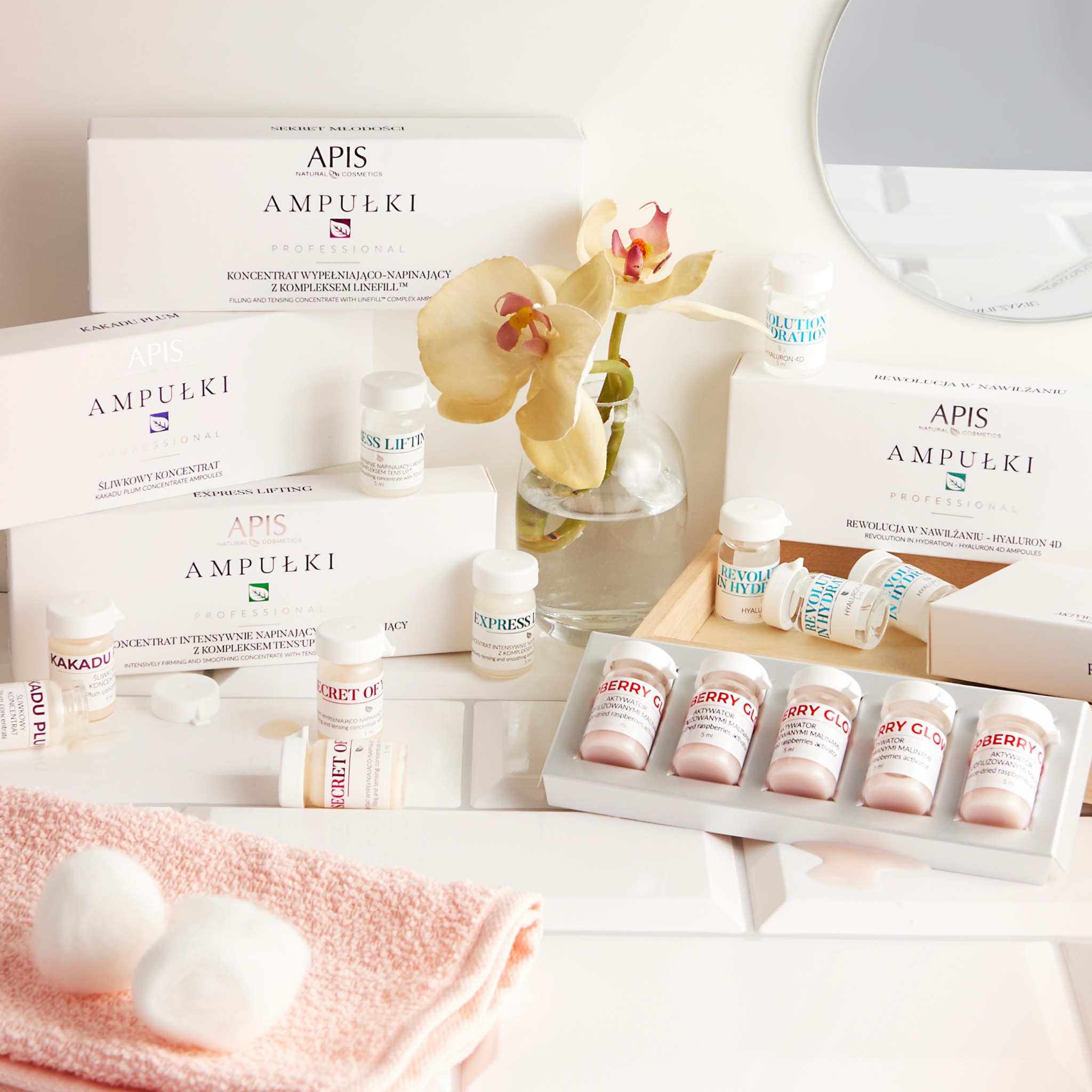 Zestaw ampułek i koncentratów marki Apis Professional, w tym serie Kakadu Plum, Express Lifting, Secret of i Berry Glow, ułożonych na białych płytkach z różowym ręcznikiem i gąbeczkami w tle...