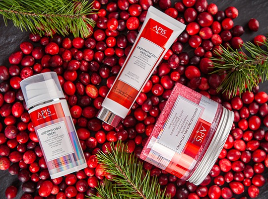 Trzy kosmetyki marki Apis: krem ujędrniający, peeling enzymatyczny i sól do kąpieli, otoczone świeżymi żurawinami i gałązką świerku na ciemnym tle.