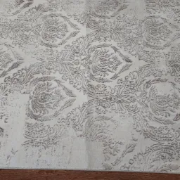 Kremowy dywan z delikatnym, wytłaczanym wzorem floralnym, leżący na ciemnobrązowej podłodze z paneli.