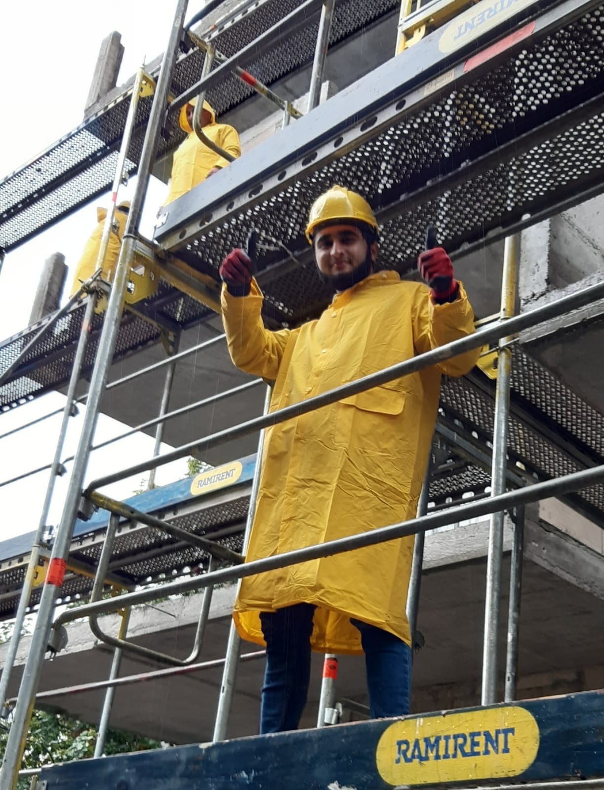 Pracownik w żółtym płaszczu i kasku na rusztowaniu w Elblągu, z podniesionymi kciukami, na tle surowej konstrukcji budynku w trakcie budowy.