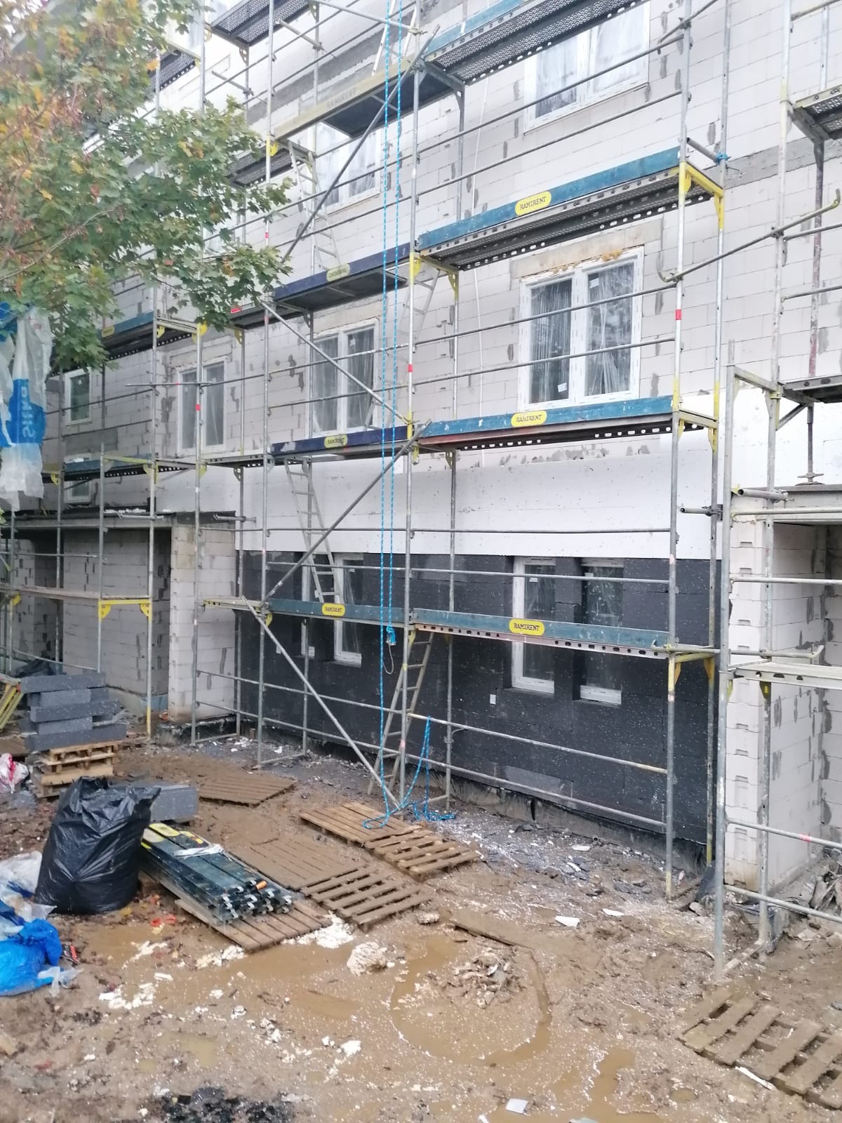 Budynek w trakcie ocieplania, widoczne rusztowania, szare bloczki, czarna izolacja, okna zabezpieczone folią, błotniste podłoże, palety i worki budowlane.