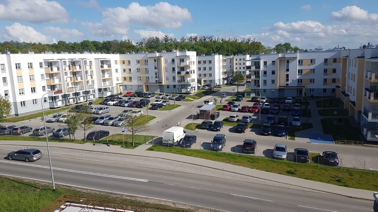 Widok z góry na osiedle z białymi budynkami wielorodzinnymi, parkingiem pełnym samochodów i dwupasmową drogą na pierwszym planie.