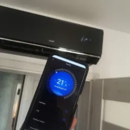 Czarny klimatyzator AUX na ścianie, sterowany smartfonem z aplikacją pokazującą temperaturę 21.5 stopni Celsjusza.