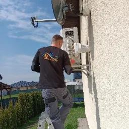 Mężczyzna na drabinie, w czarnej koszulce z logo firmy, instaluje lub serwisuje urządzenie na elewacji domu, obok anteny satelitarnej.