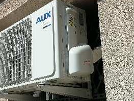 Biała jednostka zewnętrzna klimatyzatora marki AUX zamontowana na ścianie budynku o szarej, ziarnistej fakturze.