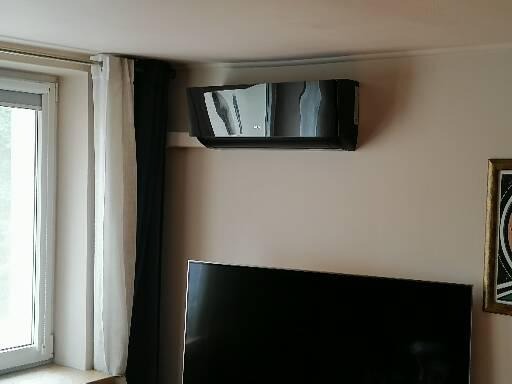 Czarny klimatyzator ścienny w salonie nad telewizorem, widok z dołu, obok okna z białymi zasłonami i fragmentu obrazu w złotej ramie.