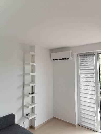 Wnętrze pokoju z zamontowanym klimatyzatorem ściennym, obok okna z żaluzjami i wąskiego regału z półkami.