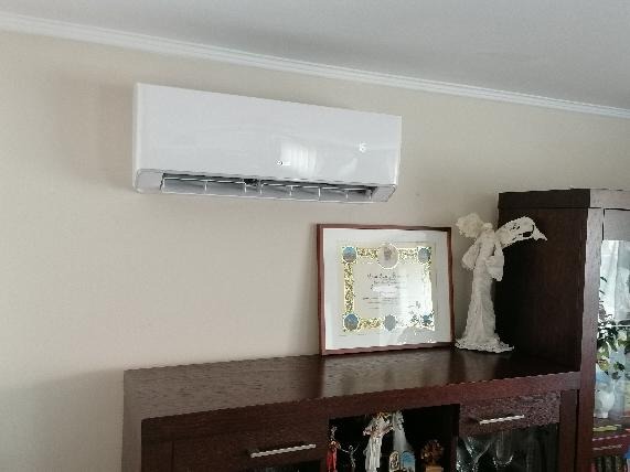 Biały klimatyzator ścienny zamontowany w pomieszczeniu nad komodą z dekoracjami, w tle biały sufit z listwą.