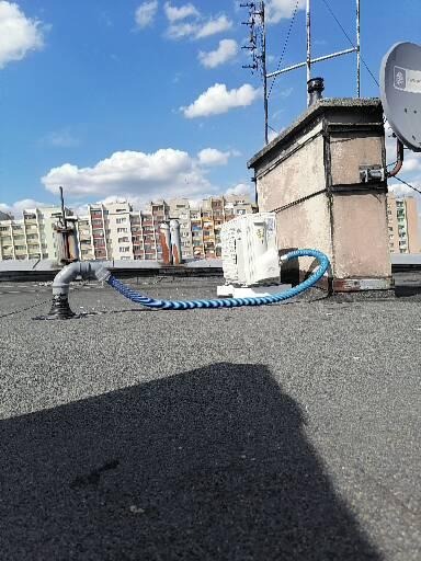 Instalacja na dachu budynku z widocznym przewodem w niebiesko-białym oplocie łączącym elementy systemu, na tle zabudowy miejskiej i błękitnego nieba z chmurami.