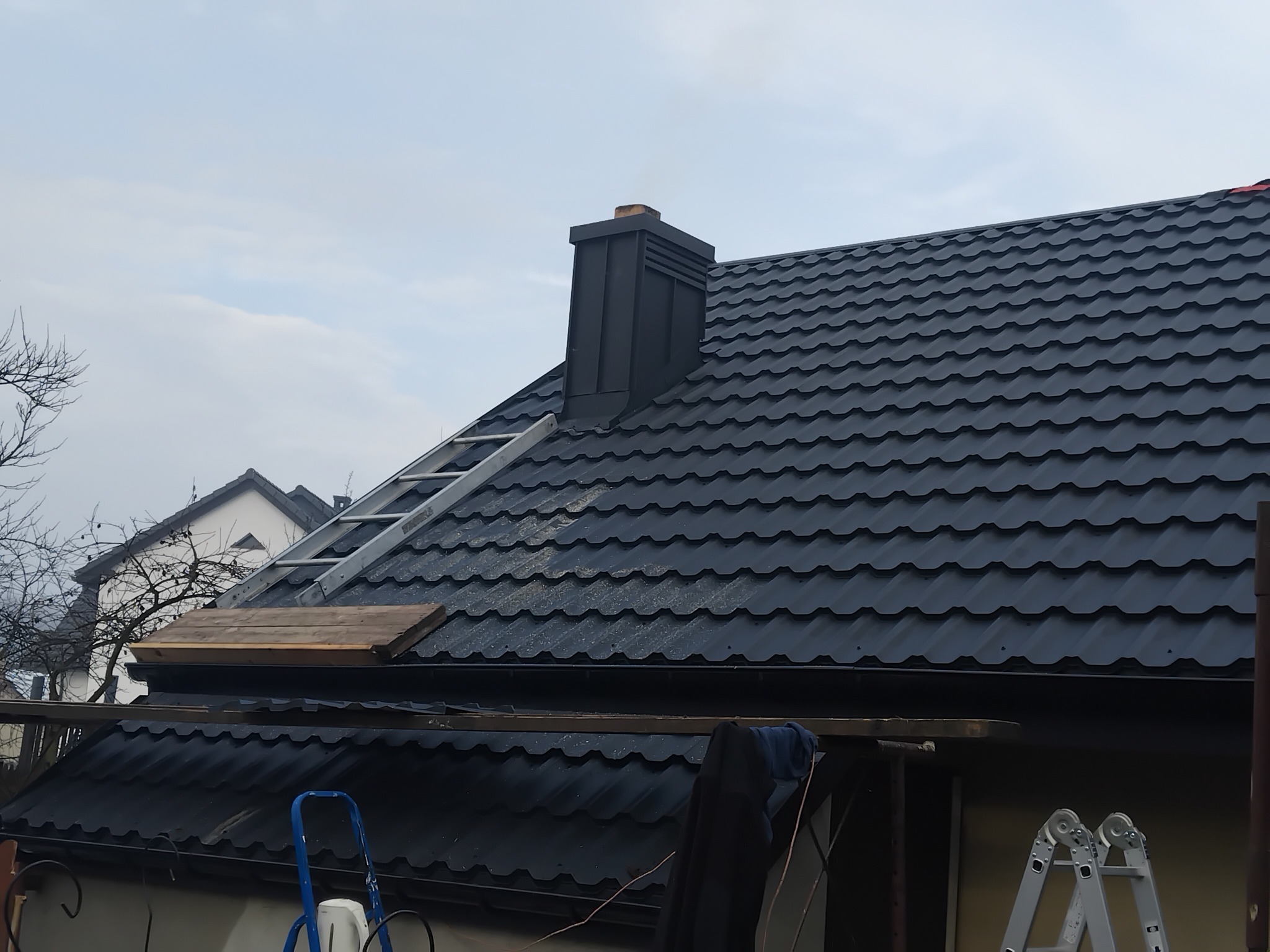 Czarny dach z blachy trapezowej z kominem i drabina oparta o dach. Widoczne narzędzia i elementy montażowe. Ujęcie z dołu, pochmurne niebo w tle.