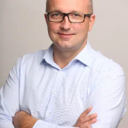 Portret mężczyzny w okularach i jasnej koszuli z założonymi rękami na neutralnym tle.